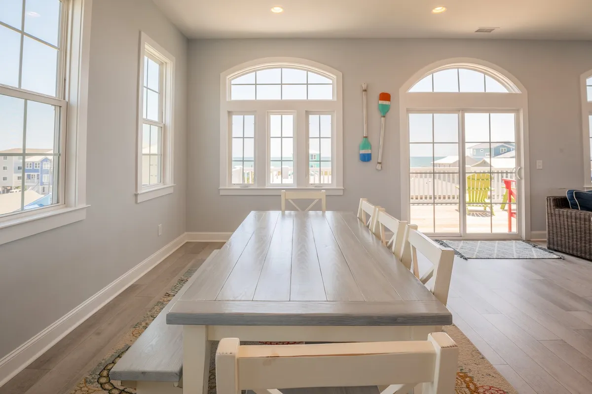 A spacious dining area featuring a large farmhouse table surrounded by chairs, with multiple windows framing outdoor views and flooding the space with light. The setup is perfect for group meals and socializing.