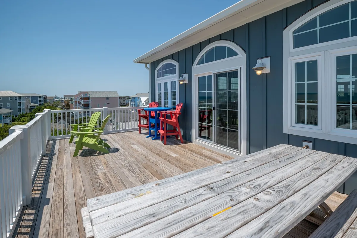 An inviting exterior deck features colorful Adirondack chairs and a wooden picnic table, set against large windows that hint at the spacious interior. The setup suggests a perfect spot for group gatherings, outdoor meals, or casual conversation while enjoying the fresh air.