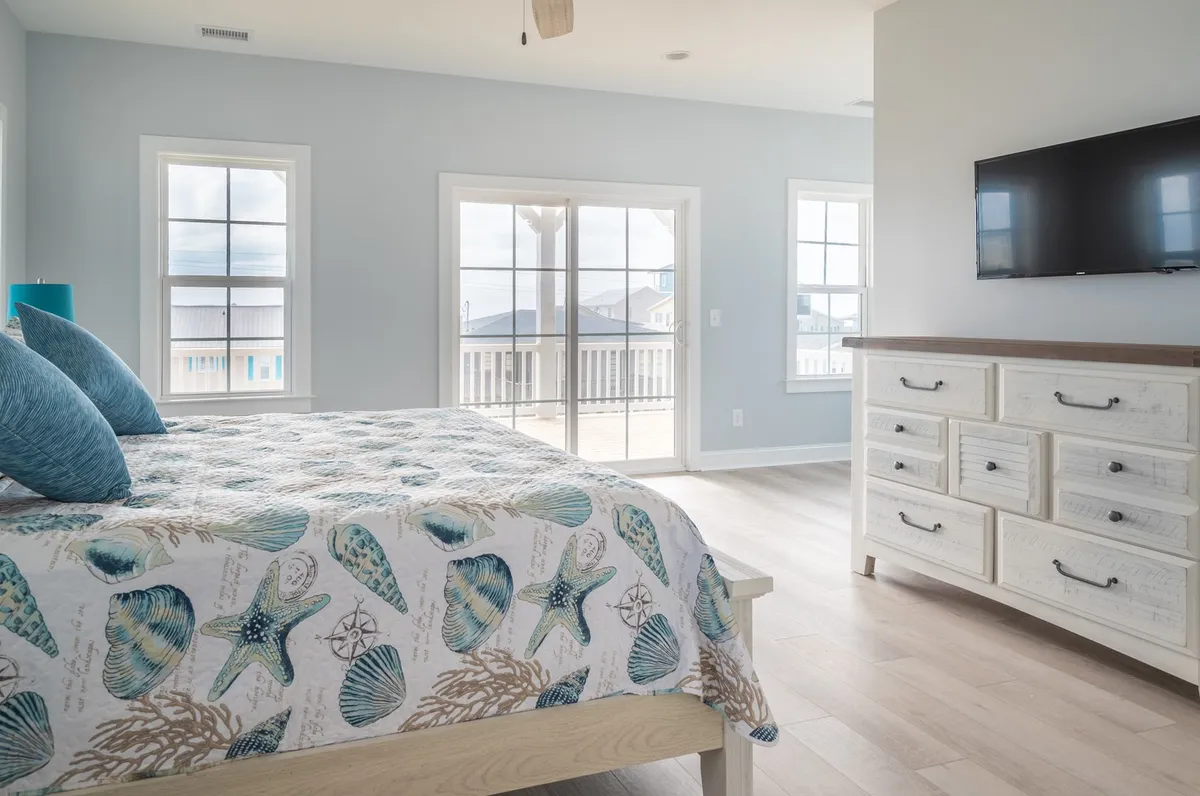A calming bedroom with seashell-patterned bedding, large windows offering a view outside, a dresser with decorative accents, and a wall-mounted TV. Morning light fills the space, creating a sense of tranquility and comfort. A private door suggests access to an outdoor deck, in line with suite features described.