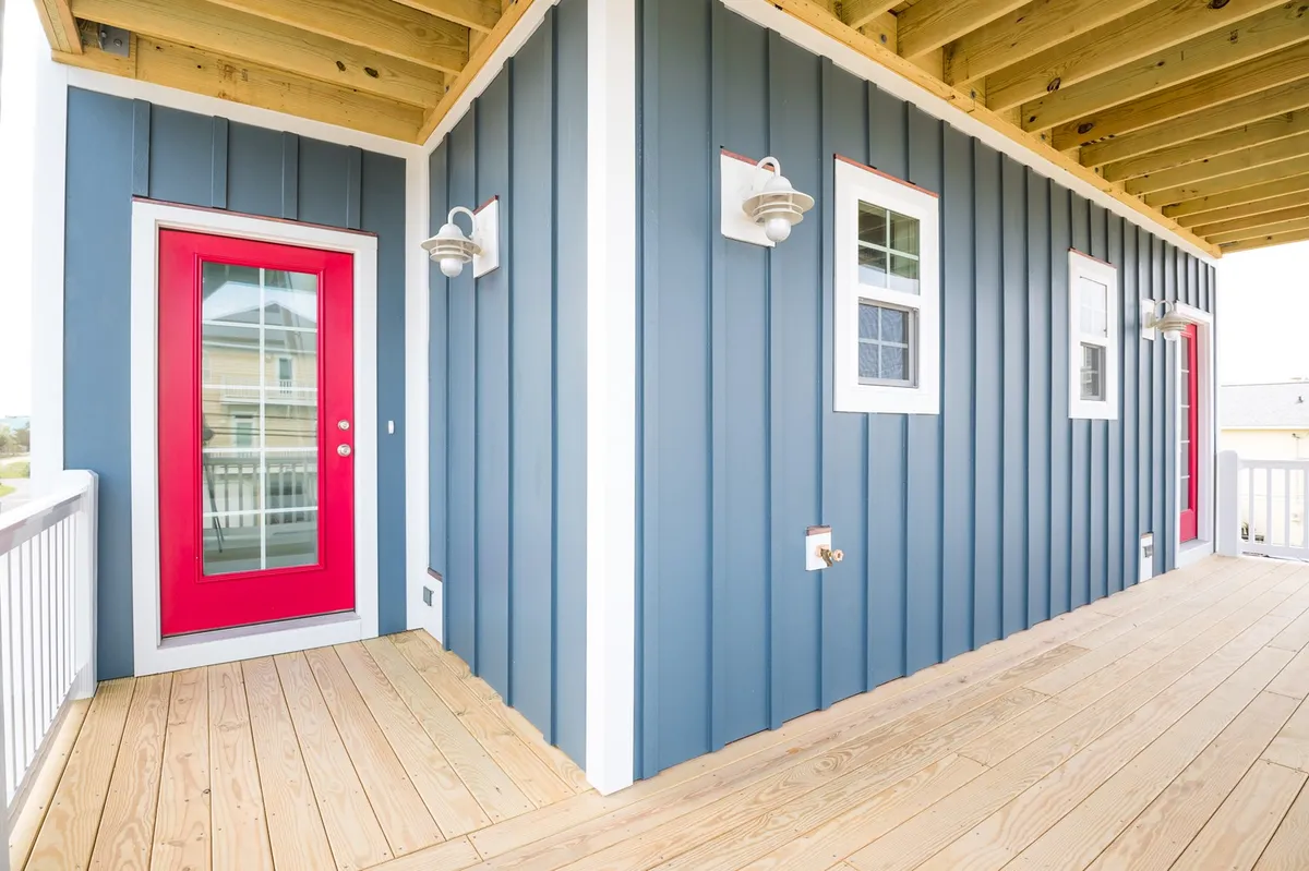This image showcases the exterior of Patriots Place, highlighting a cheerful red door set into blue wooden siding with white trim and outdoor lighting fixtures. The porch is arranged for relaxing and socializing, suggesting an inviting entryway after a beach day and a welcoming environment for guests and pets alike.