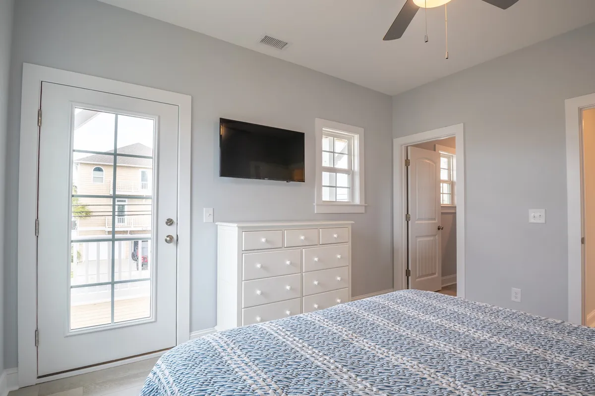A tranquil bedroom featuring patterned bedding, a white dresser, a television mounted on the wall, and a door that leads to an outdoor space. Soft natural light filters in, and the overall mood is peaceful and inviting.