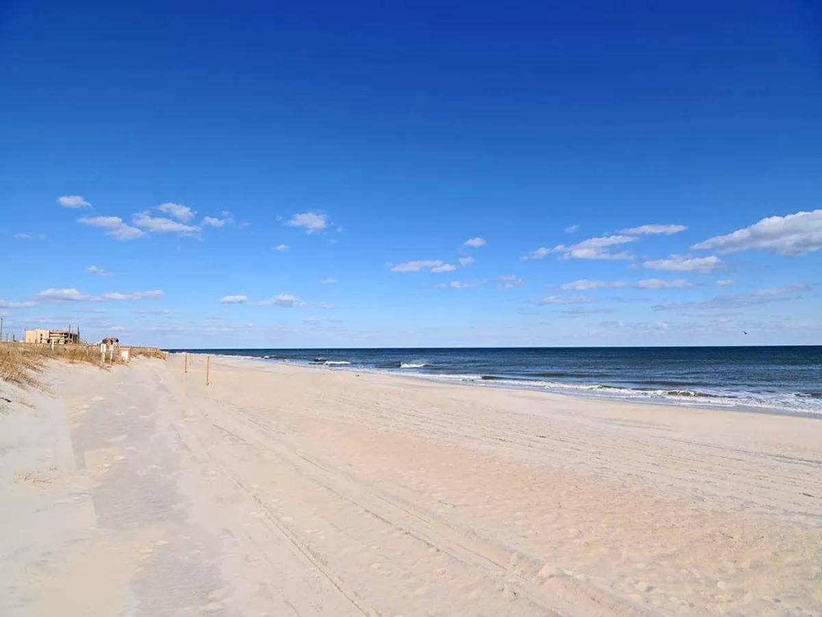 A wide, inviting stretch of sandy beach with gentle ocean waves under a clear blue sky. The shoreline is perfect for sunbathing, swimming, and building sandcastles, with a few distant beachgoers adding a sense of vibrant activity. Distant buildings hint at nearby attractions and easy access from the property.