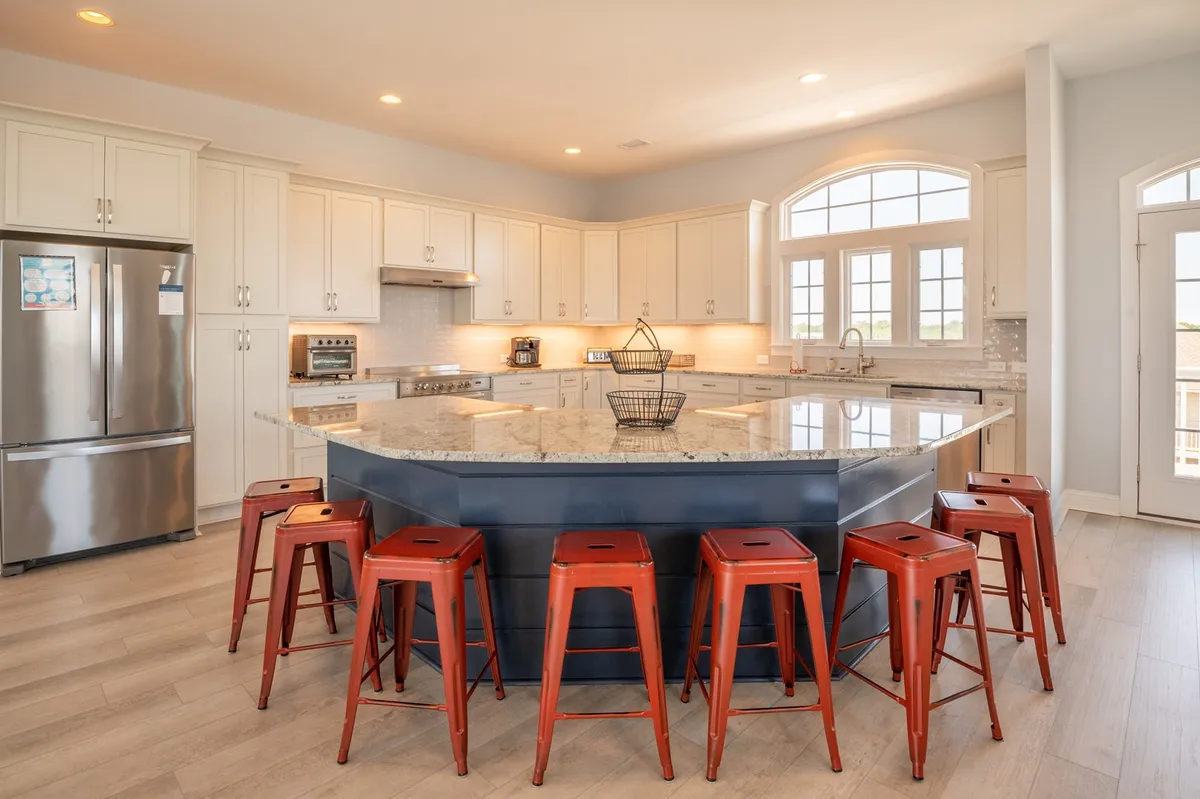 A bright, inviting kitchen with a large island and red metal stools, bathed in natural light from surrounding windows. The kitchen is equipped with luxury appliances and features space for both cooking and casual dining.