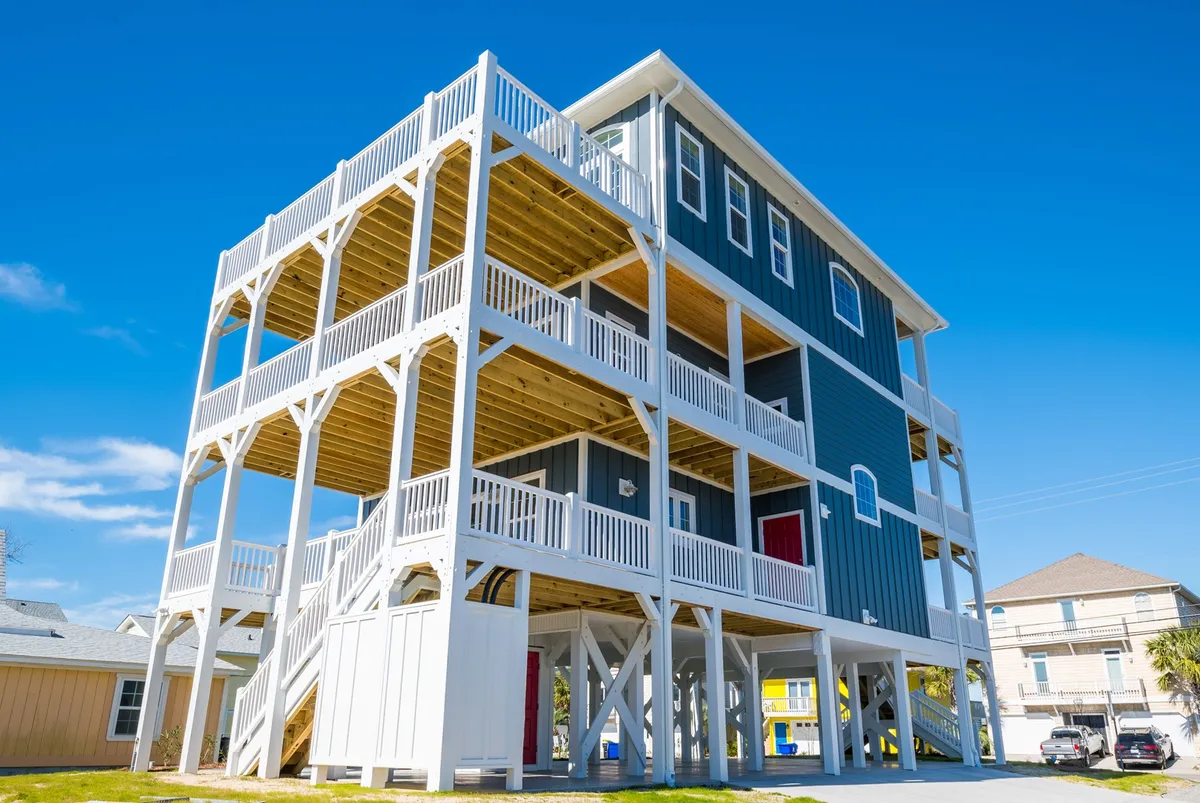 A vibrant exterior view of the stilted beach house, showcasing multiple balconies with colorful doors. The image captures the expansiveness of the property and the inviting decks designed for sunbathing, relaxing, and taking in the surrounding coastal scenery.