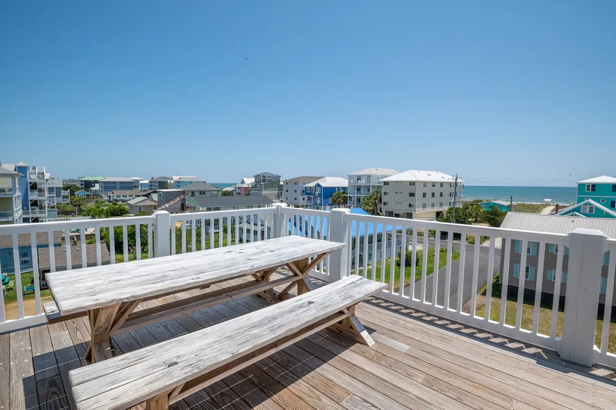 The image features a wooden picnic table on an exterior deck, with an ocean view and nearby houses in sight. The setting is casual and inviting, suggesting outdoor dining, sunset dinners, or morning coffee while enjoying the sea breeze with pets or family members.
