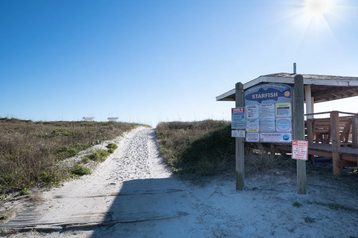 An exterior scene showing a signpost, a sandy pathway leading towards the beach, and a small seating area. The mood is tranquil, evoking the anticipation of setting out for adventure or returning from a coastal excursion.