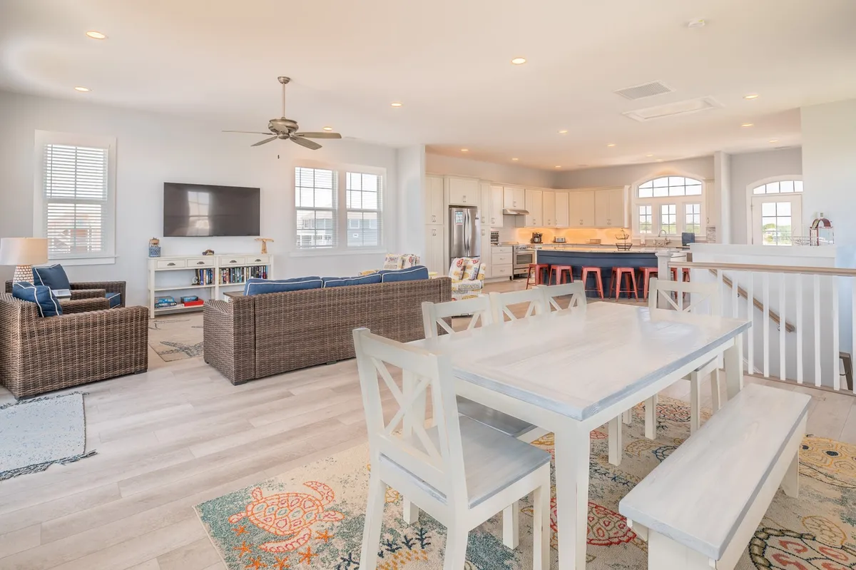 This image captures an expansive open-concept interior, featuring a plush living area with ample seating, a modern farmhouse-style dining table, and a glimpse of the kitchen area. A large TV is visible, and the space is filled with natural light from generous windows.